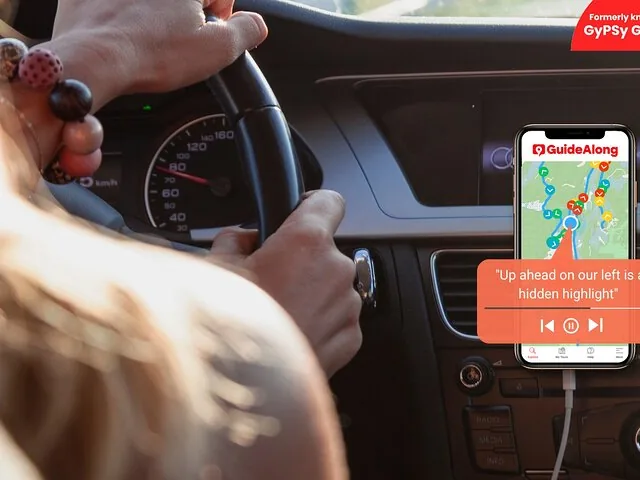 Persona alla guida di un'auto con l'app per audioguide su smartphone visibile.